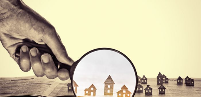 Hand holds the magnifying glass in front of an open newspaper with paper houses. That could mean rent, search, purchase real estate.